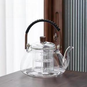 以下が含まれることがあります： 曲がった注ぎ口と内蔵のインフューザーを備えた透明なガラスティーポット。木製の蓋と黒と茶色のハンドルが付いています。 ティーポットは、お茶を淹れるように設計されています。