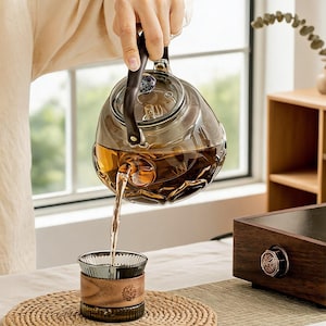 May include: A smoke-tinted glass teapot with a dark handle and a textured, faceted base, pouring amber-colored tea into a matching smoke-tinted glass cup that features a wide, smooth wooden band around its center and a ribbed glass base.