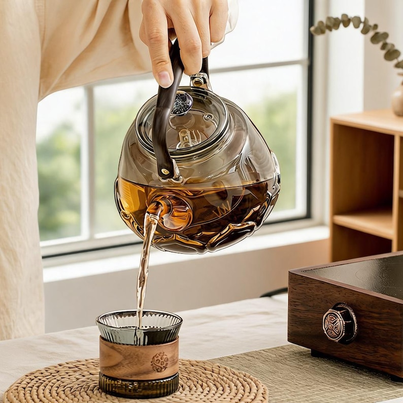 May include: A smoke-tinted glass teapot with a dark handle and a textured, faceted base, pouring amber-colored tea into a matching smoke-tinted glass cup that features a wide, smooth wooden band around its center and a ribbed glass base.