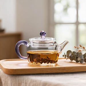 May include: A clear glass teapot with a purple-tinted handle and lid knob. The teapot has a wide, cylindrical body filled with amber-coloured tea and loose tea leaves. It features a short, angled spout and a flat, circular lid with a small handle.