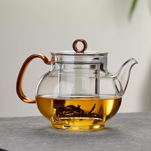 May include: A clear glass teapot with a brown handle and lid detail. The teapot is filled with amber-coloured liquid and tea leaves. The design includes a built-in infuser for steeping tea. The teapot is sitting on a grey surface.