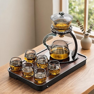 May include: A tea set featuring a dark-tinted glass teapot with a matching lid and a suspended brewing chamber, accompanied by six small, rounded glass cups filled with amber liquid, all arranged on a rectangular dark wooden serving tray.
