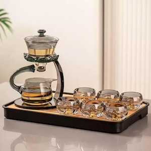 May include: A glass tea set with a teapot, infuser, and six small cups. The set has gold accents and sits on a black tray with a bamboo base. The teapot has a curved handle and a spout.