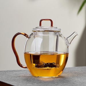 May include: A clear glass teapot with a brown handle and lid handle. The teapot is filled with amber-colored tea and has a built-in infuser. The teapot is sitting on a gray surface.