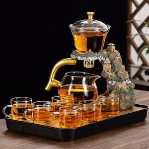 以下が含まれることがあります： 透明なガラスのティーポット、ガラスのインフューザー、6つの小さなティーカップが入ったティーセット。すべてお茶で満たされています。セットには、ミニチュアの建物と金色のハンドルが付いた装飾的な山型のスタンドが含まれています。