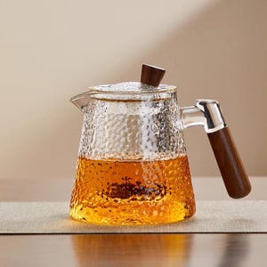 May include: A clear glass teapot with a textured surface, filled with amber liquid. It features a wooden handle and lid knob. The teapot is designed for brewing tea and sits on a neutral-coloured surface.