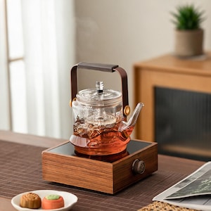 Peut inclure: Une théière en verre transparent avec une anse marron foncé et un couvercle assorti, remplie de thé ambré. La théière repose sur une base carrée en bois dotée d'un bouton de commande circulaire sur le devant, conçue pour chauffer le thé.
