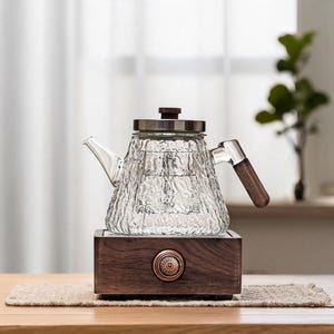 May include: A clear glass teapot with a textured, rippled surface, featuring a dark wood handle and a matching wooden lid knob. The teapot rests on a rectangular dark wood base with a central circular copper-toned power button and metallic trim.