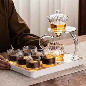 May include: A clear glass tea set with a teapot, six small glasses with wooden bands, and a tea server. The tea set is on a white tray. The tea is amber-coloured. The tea server has a clear glass lid.