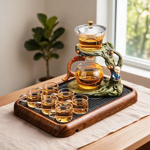 Peut inclure: Un service à thé en verre comprenant une théière transparente et six petites tasses en verre disposées sur un plateau rectangulaire en bois. L'ensemble inclut un support décoratif en forme d'arbre vert avec une figurine de moine.