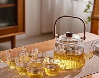 May include: A glass tea set comprising a teapot and six teacups. The teapot features a wooden handle and lid. The teacups have handles and are filled with tea. The set is displayed on a wooden table.