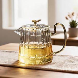 May include: A clear glass teapot with vertical ribbed detailing, a gold-colored handle, and a matching gold-colored lid featuring a small decorative finial. The teapot contains a light yellow liquid and has a wide, flat base with a starburst pattern.