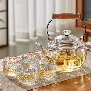 Puede incluir: Una tetera de vidrio transparente con un asa de madera y cuatro vasos texturizados a juego. La tetera contiene té y un colador con flores secas. Los vasos están llenos de té, creando un juego de té cálido.