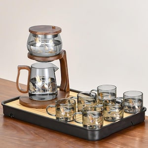 May include: A glass tea set with a wooden stand and tray. The set includes a teapot, a brewing chamber, and six teacups. Each glass piece features a gold crane design. The wooden stand and tray have a dark brown finish.