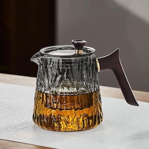 May include: A clear glass teapot with a textured surface and a dark wooden handle. The teapot has a dark wooden lid with a gold accent. The teapot is filled with a golden-brown liquid, possibly tea.