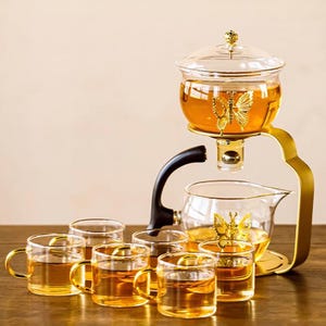 May include: A glass tea set with a gold butterfly design. The set includes a brewing pot, a serving carafe, and several small teacups. The tea is amber-coloured, and the set has a gold-coloured metal stand.