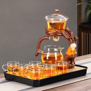 May include: A tea set featuring a clear glass teapot, cups, and a serving vessel, all filled with tea. The set includes a wooden stand with a small statue and a black tray. The tea set is designed for serving tea.