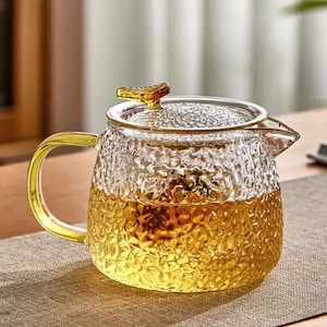 May include: A clear glass teapot with a textured surface and a golden handle and lid. The teapot is filled with a golden liquid, likely tea, and sits on a neutral-colored surface. The design is elegant and suitable for serving tea.