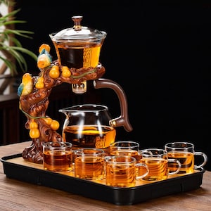 Può includere: Servizio da tè decorativo con una teiera in vetro, un infusore in vetro e sei piccole tazze di vetro. Il set include un supporto in legno con un design a ramo intagliato, con due figurine di uccelli colorati e diversi frutti arancioni. Il tè è color ambra.