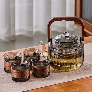 May include: A glass tea set comprising a teapot and four glasses. The teapot features a wooden handle and a clear, ridged glass body. The glasses are encircled by wooden bands. The set is displayed on a neutral-toned table runner.
