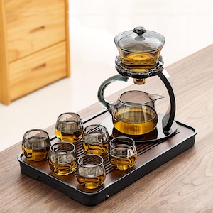 Può includere: Un moderno set da tè con una teiera in vetro, un infusore e sei bicchieri piccoli, tutti riempiti di tè. Il set è posizionato su un vassoio di legno scuro, con un supporto in metallo nero che sostiene l'infusore e la teiera. Il tè è di colore ambra dorato.