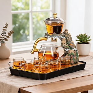 May include: A glass tea set featuring a clear teapot and six small glass cups filled with amber tea. The set includes a decorative mountain-shaped stand with a gold-colored handle and a black rectangular serving tray for the tea service.