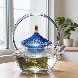May include: A clear glass teapot featuring a textured body and a vibrant blue, pagoda-style lid topped with a small amber-coloured finial. The teapot has a tall, arched glass handle and a short spout, containing loose green tea leaves in water.