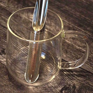 May include: A clear glass mug with a handle holds a stainless steel tea infuser. The infuser is cylindrical with small holes and a scoop at the top. The mug sits on a wooden surface.