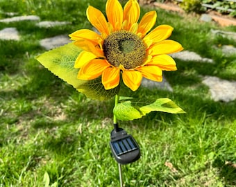 Könnte beinhalten: Eine künstliche gelbe Sonnenblume mit grünen Blättern, die an einem schwarzen solarbetriebenen Lichtpfahl befestigt sind. Der Pfahl ist in den Boden gesteckt.