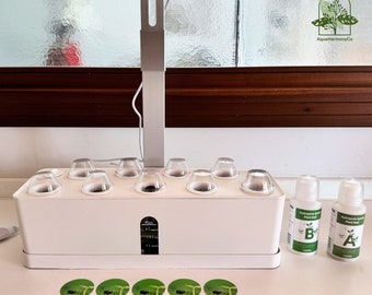 Op de afbeelding: Wit hydroponisch tuinsysteem met negen doorzichtige plastic plantpotten, een zilveren groeilamp en twee flessen plantenvoeding met de labels "A" en "B".