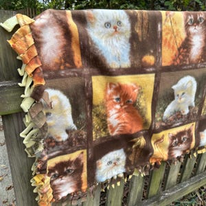 Op de afbeelding: Een fleecedeken met een herhaald patroon van schattige kittens in verschillende kleuren en poses. De deken heeft een bruine rasterachtergrond met gele en oranje accenten. De randen zijn afgewerkt met een franjebies.