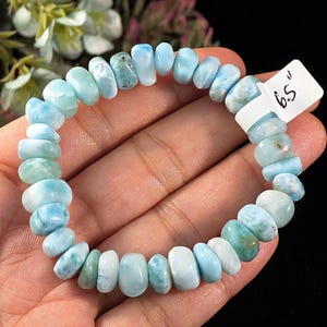 Puede incluir: Una pulsera el&aacute;stica compuesta por cuentas de Larimar redondas y pulidas en varios tonos de azul claro, turquesa y blanco. Una etiqueta blanca unida a la pulsera muestra el texto 6.5 inches, indicando la circunferencia total de la pieza.