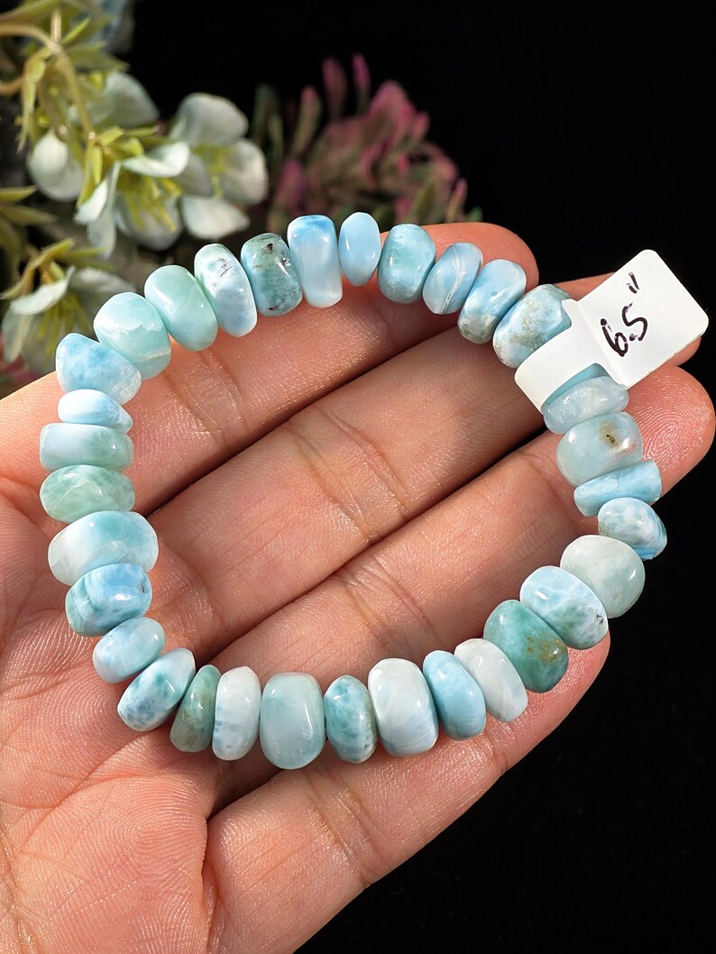 Puede incluir: Una pulsera el&aacute;stica compuesta por cuentas de Larimar redondas y pulidas en varios tonos de azul claro, turquesa y blanco. Una etiqueta blanca unida a la pulsera muestra el texto 6.5 inches, indicando la circunferencia total de la pieza.