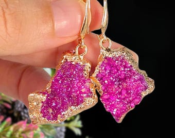 May include: A pair of gold-colored earrings featuring irregular, magenta-colored crystal formations. The crystals are encased in a gold-colored metal setting, creating a striking contrast. The earrings have a hook closure.