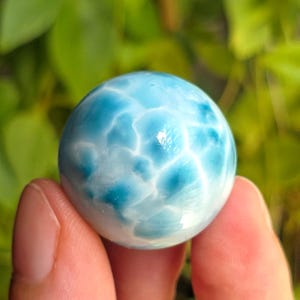 Puede incluir: Una gema esférica pulida, principalmente azul claro con vetas blancas y azul oscuro. La piedra se sostiene entre los dedos, con un fondo verde borroso de follaje.