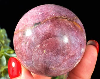 May include: A polished spherical gemstone in shades of deep magenta and dusty rose, featuring a prominent horizontal fissure with dark brown and golden mineral inclusions running across the center of the smooth, rounded surface.
