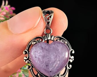 May include: A heart-shaped pendant with a light purple stone set in an ornate, silver-toned frame. The pendant has a decorative bail at the top for a chain. The frame features scrolling filigree designs. The background is dark.