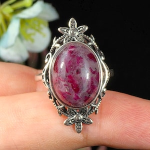 Op de afbeelding: Een zilverkleurige ring met een ovaalvormige cabochonsteen met diep magenta en grijze gevlekte patronen. De metalen zetting is gedetailleerd met ingewikkeld filigraanwerk en kleine bloemmotieven aan de boven- en onderkant van de steen.