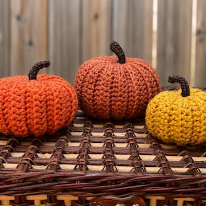 May include: Three crocheted pumpkins in varying sizes and colours. Two pumpkins are shades of orange, and one is mustard yellow. Each pumpkin features a dark brown crocheted stem attached to the top, with a ribbed texture throughout the body.