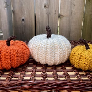 May include: Three crocheted pumpkins in orange, white, and yellow, each featuring a dark brown stem. The pumpkins are crafted with a ribbed texture and are arranged in a row, showcasing their rounded shapes and distinct yarn colours.