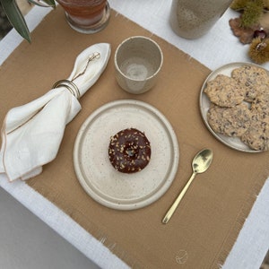 Peut inclure: Une assiette en céramique beige et brune mouchetée avec un beignet glacé au chocolat surmonté de noix hachées. L'assiette est sur un set de table brun avec une serviette en lin blanche et une cuillère dorée.