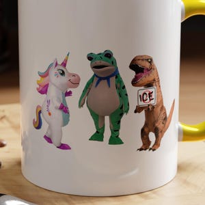 May include: White ceramic mug with a yellow interior and handle. The mug features a whimsical design of a unicorn, frog, and dinosaur. The dinosaur holds a sign that says "ICE". The mug sits on a wooden surface.
