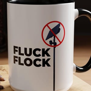 May include: A white ceramic mug with a black interior and handle. The exterior features the black text FLUCK FLOCK stacked vertically and a graphic of a black satellite dish on a pole crossed out by a red circle. The design is printed on the front.