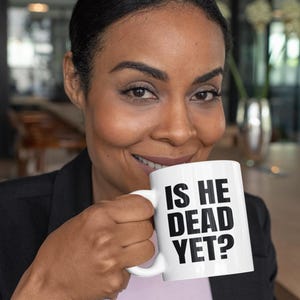 May include: A white ceramic mug with a black inscription that reads "Is he dead yet?" A person is holding the mug with their right hand.
