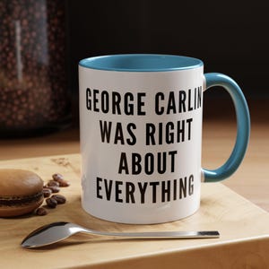 Puede incluir: Taza de cerámica blanca con interior y asa azules. La taza presenta el texto negro "GEORGE CARLIN WAS RIGHT ABOUT EVERYTHING". Una cuchara y un macaron están sobre una superficie de madera.