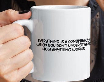 May include: White ceramic mug with the text "EVERYTHING IS A CONSPIRACY WHEN YOU DON'T UNDERSTAND HOW ANYTHING WORKS" in black. The mug is held by a person with a black manicure.
