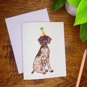 May include: Greeting card featuring a watercolor illustration of a German Shorthaired Pointer wearing a party hat. The dog is brown and white with a black nose and floppy ears. The party hat is yellow and green. A blank white envelope is next to the card.
