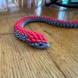 Puede incluir: Un juguete de serpiente articulada impreso en 3D en rojo y gris. La cabeza y la espalda de la serpiente son rojas, mientras que el cuerpo es gris. El juguete está sobre una superficie de madera.