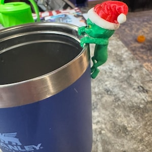 May include: A green Grinch figure wearing a red and white Santa hat is perched on the rim of a blue insulated tumbler. The tumbler has a silver rim and the word "STANLEY" is visible on the side.