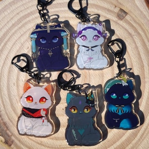 Puede incluir: Cinco llaveros con forma de gato con herrajes negros. Cada gato tiene un diseño único, incluyendo diferentes colores y accesorios. Los gatos son en tonos de blanco, rojo, azul y negro. Los llaveros están sobre una superficie de madera.